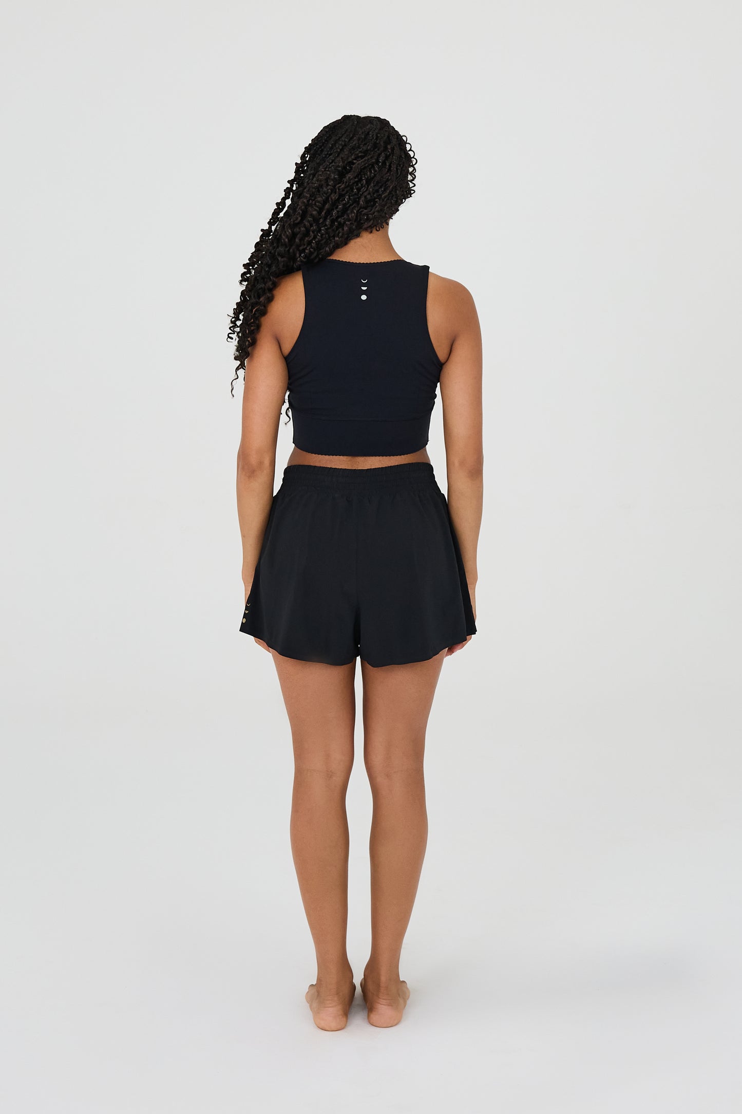 back view of model wearing L’Original Flowy Short featuring a featherlight pleated outer layer and a sculpting compressive inner short. The 2-in-1 design blends feminine movement with performance support, including patented MicroPerle® bead infusion and a hidden inner pocket for essentials like a phone or tennis ball.