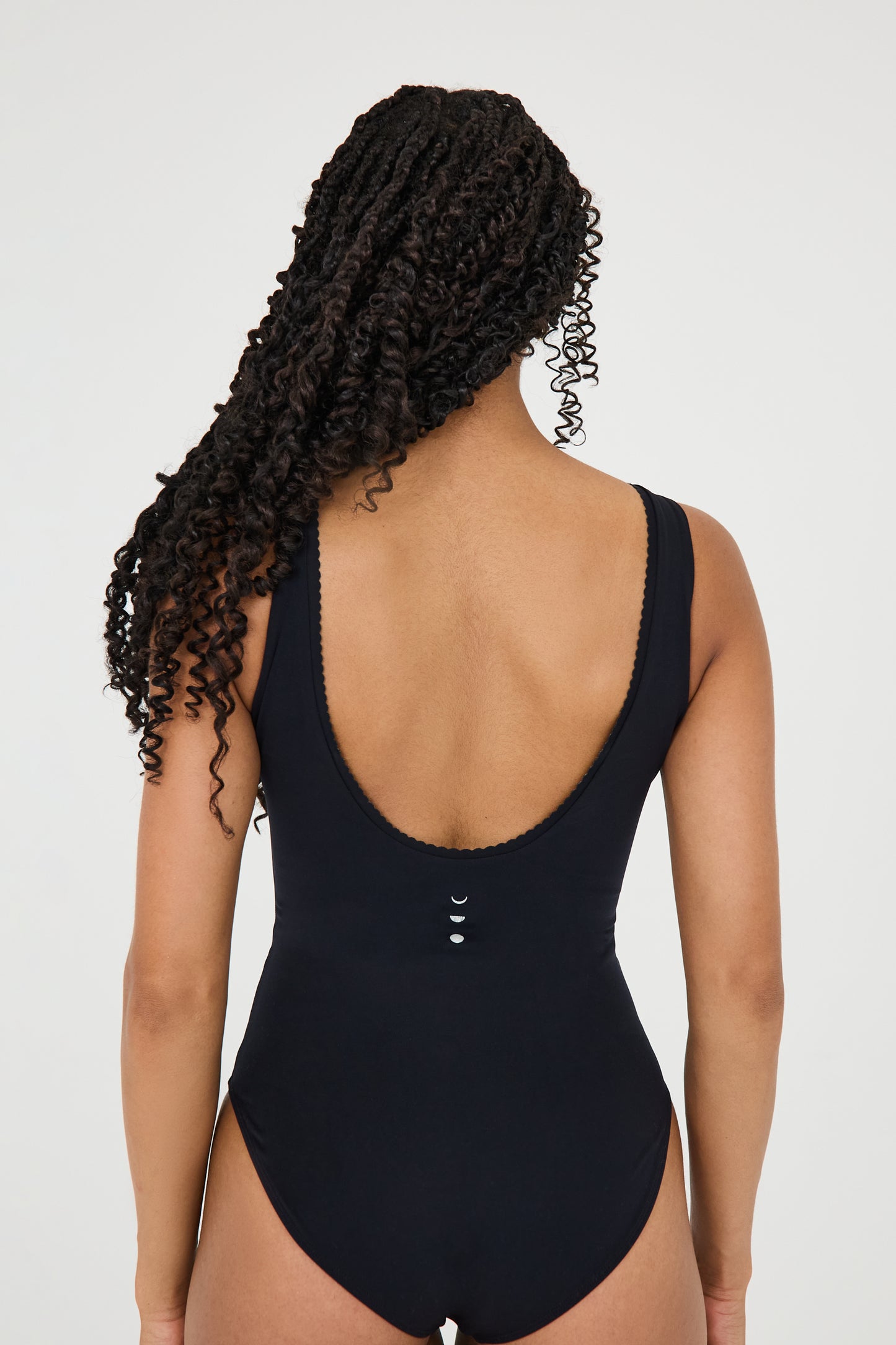 Back view of model wearing the black Adorn Bodysuit from Elastique’s sculptural Adorn Collection, featuring a flattering décolleté neckline with scalloped trim, open back design, smooth, form-fitting silhouette designed for both swim and everyday wear. Made with UPF 50 fabric and infused with patented MicroPerle® beads for gentle lymphatic stimulation and core support