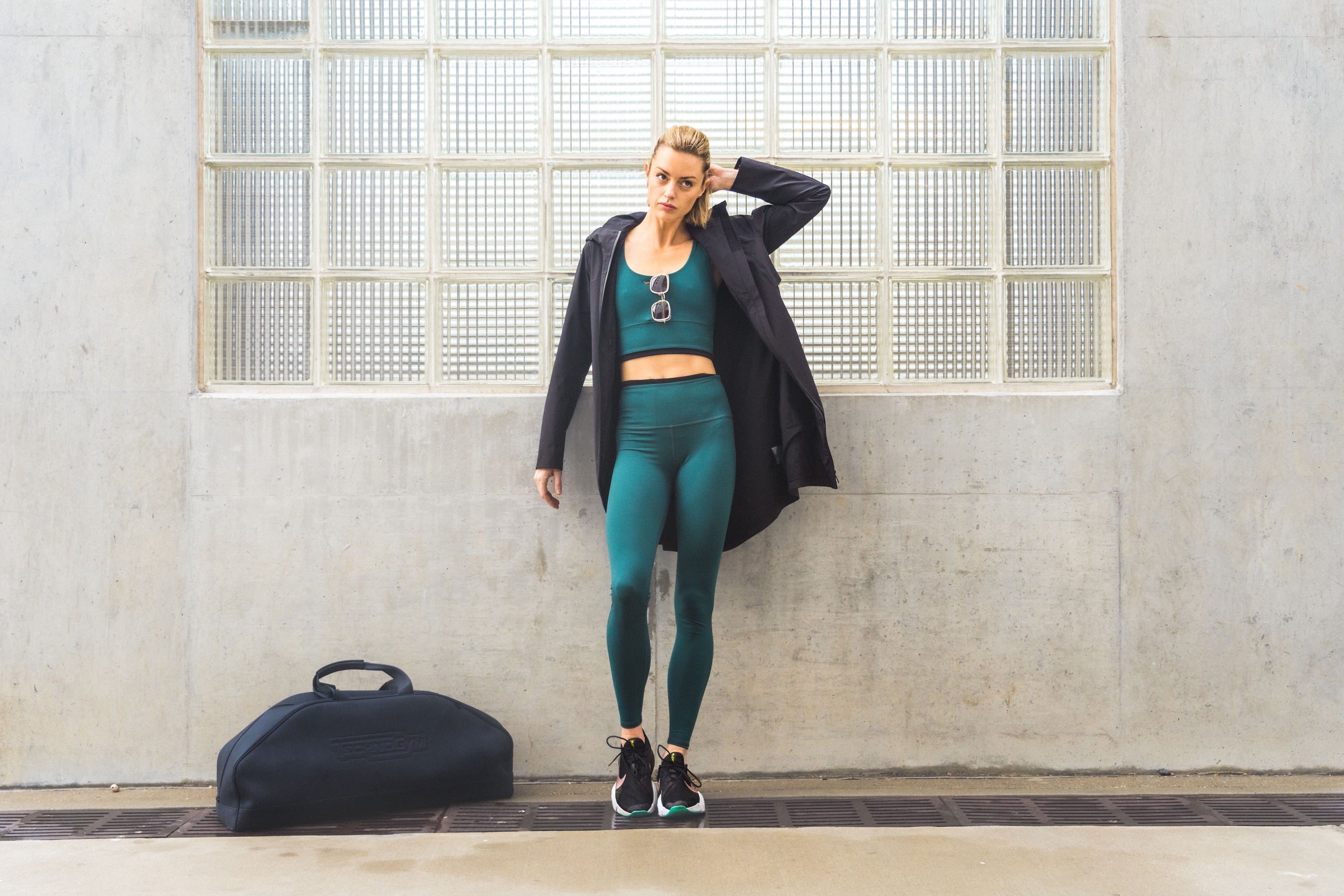 Women leaning against concrete and glass wall in green leggings and bra top. Black jacket, sunglasses hooked into top of bra top with travel bag on the ground.