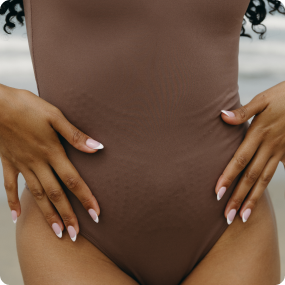 Model wearing the black Adorn Bodysuit from Elastique’s sculptural Adorn Collection, featuring a flattering décolleté neckline with scalloped trim, open-back design, and a smooth, form-fitting silhouette. Crafted with UPF 50 OEKO-TEX® fabric infused with patented MicroPerle® beads for gentle lymphatic stimulation, improved circulation, and core support.