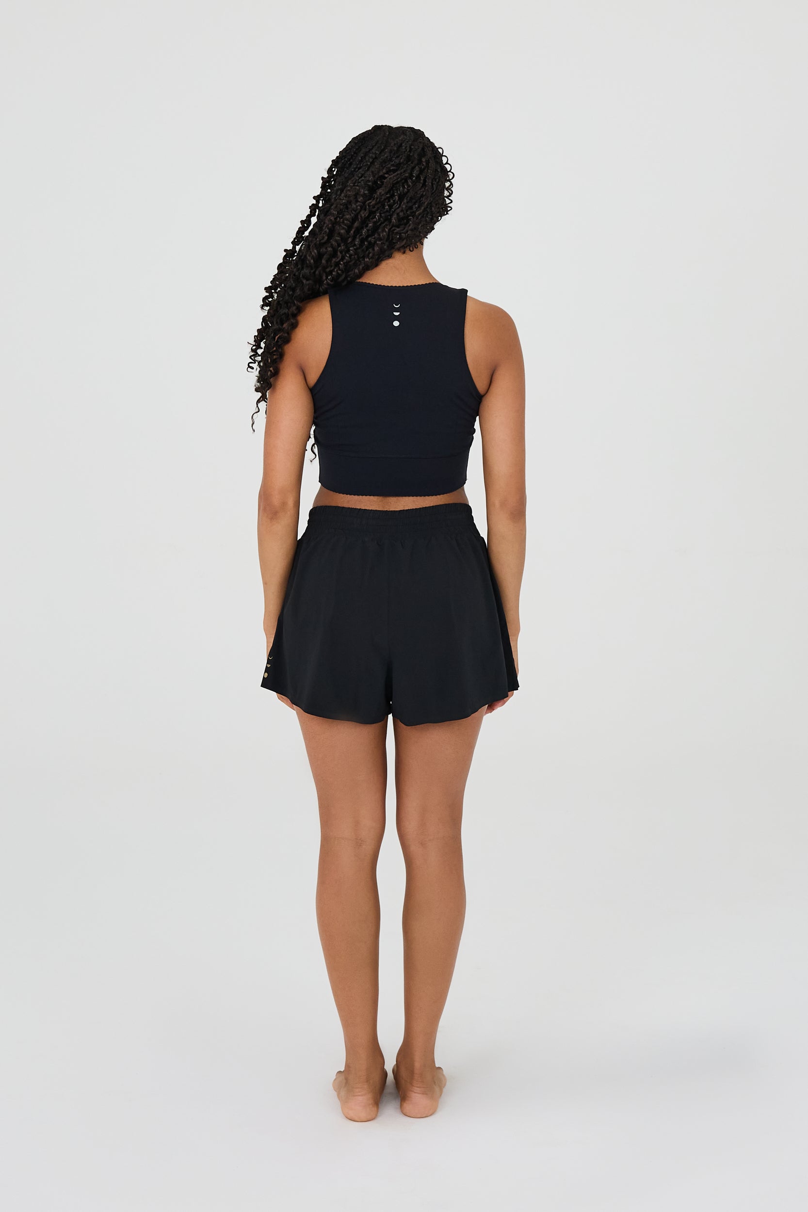 back view of model wearing L’Original Flowy Short featuring a featherlight pleated outer layer and a sculpting compressive inner short. The 2-in-1 design blends feminine movement with performance support, including patented MicroPerle® bead infusion and a hidden inner pocket for essentials like a phone or tennis ball.