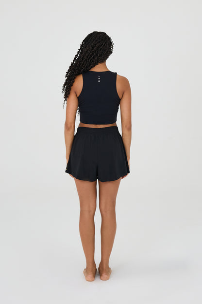 back view of model wearing L’Original Flowy Short featuring a featherlight pleated outer layer and a sculpting compressive inner short. The 2-in-1 design blends feminine movement with performance support, including patented MicroPerle® bead infusion and a hidden inner pocket for essentials like a phone or tennis ball.
