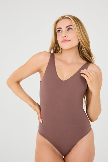 Model wearing the cappuccino Adorn Bodysuit from Elastique’s sculptural Adorn Collection, featuring a flattering décolleté neckline with scalloped trim, open back design, smooth, form-fitting silhouette designed for both swim and everyday wear. Made with UPF 50 fabric and infused with patented MicroPerle® beads for gentle lymphatic stimulation and core support