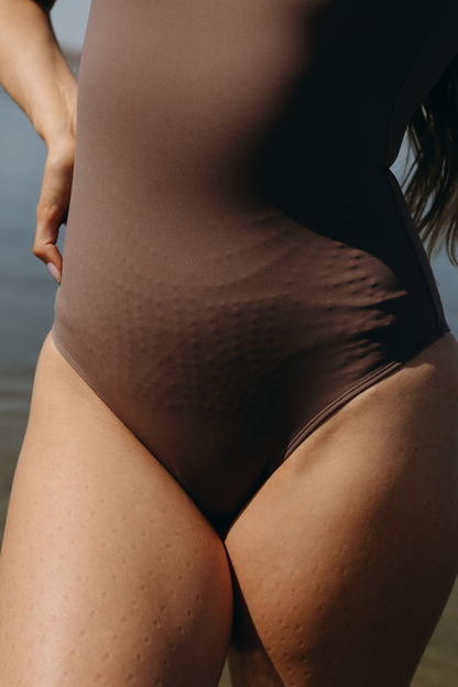 Model wearing the cappuccino Adorn Bodysuit from Elastique’s sculptural Adorn Collection, featuring a flattering décolleté neckline with scalloped trim, open back design, smooth, form-fitting silhouette designed for both swim and everyday wear. Made with UPF 50 fabric and infused with patented MicroPerle® beads for gentle lymphatic stimulation and core support
