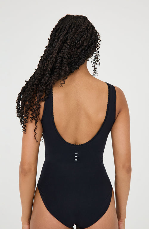 Load image 1 in gallery view. Model wearing the black color Adorn Bodysuit from Elastique’s sculptural Adorn Collection, featuring a flattering décolleté neckline with scalloped trim, open-back design, and a smooth, form-fitting silhouette. Crafted with UPF 50 OEKO-TEX® fabric infused with patented MicroPerle® beads for gentle lymphatic stimulation, improved circulation, and core support.