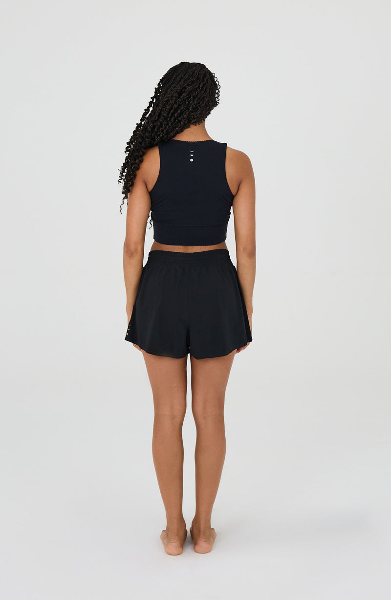 Model wearing the L’Original Flowy Short in black with a featherlight pleated outer layer and sculpting compressive inner short. This 2-in-1 design combines feminine movement with performance support, featuring Elastique’s patented MicroPerle® bead infusion and a hidden inner pocket for essentials like a phone or tennis ball.