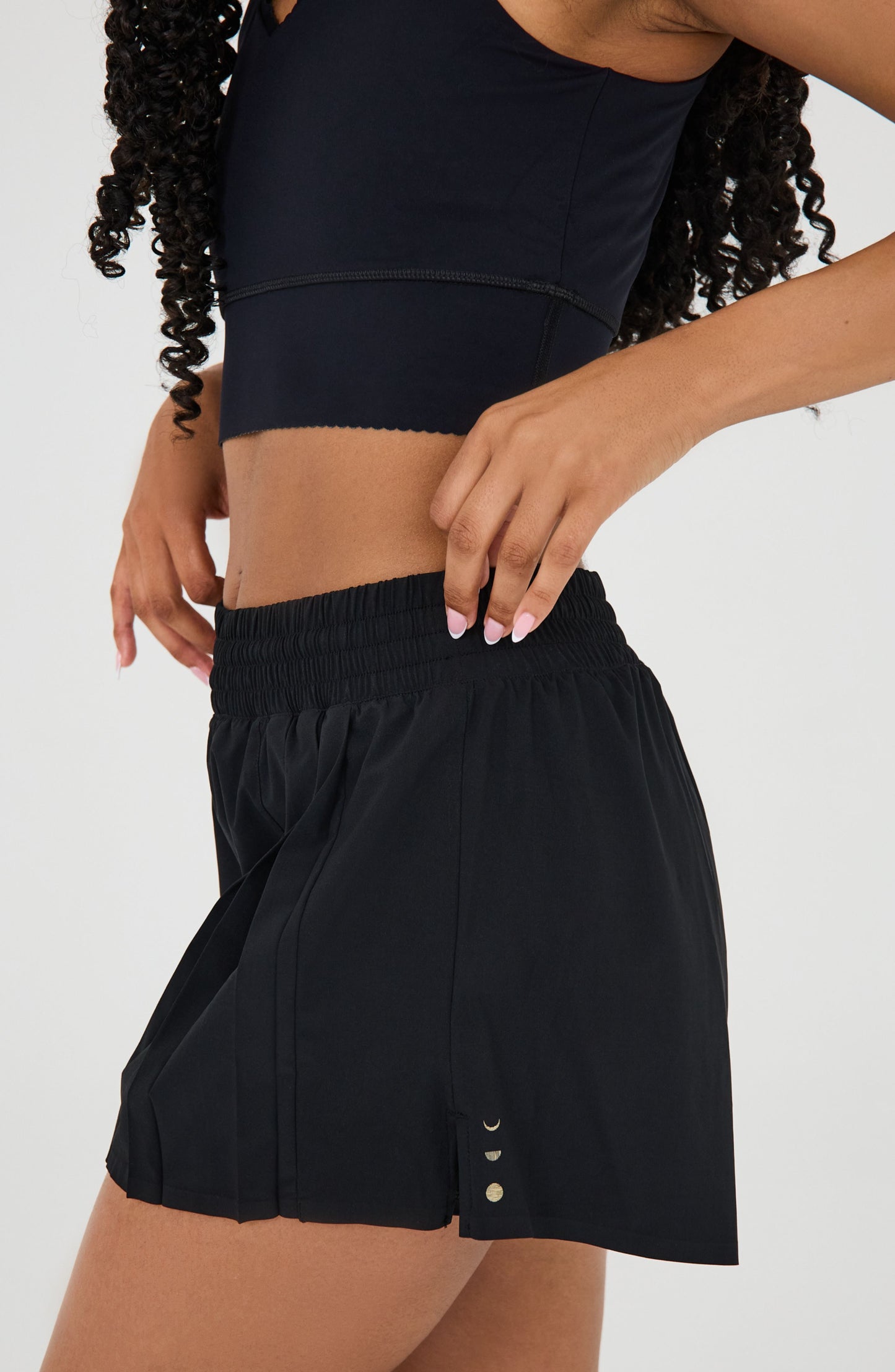 Model wearing the L’Original Flowy Short in black with a featherlight pleated outer layer and sculpting compressive inner short. This 2-in-1 design combines feminine movement with performance support, featuring Elastique’s patented MicroPerle® bead infusion and a hidden inner pocket for essentials like a phone or tennis ball.