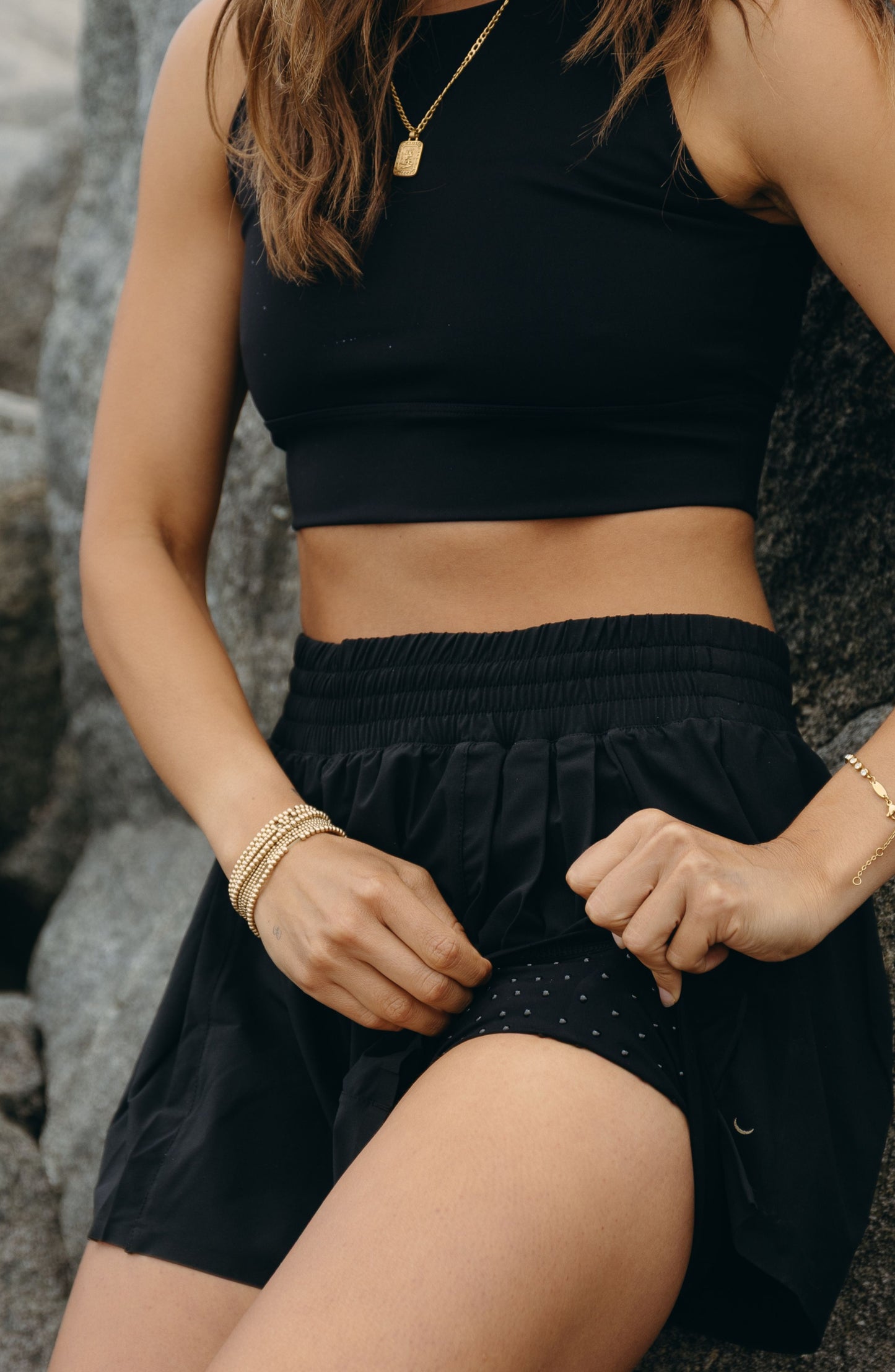 Model wearing the L’Original Flowy Short in black with a featherlight pleated outer layer and sculpting compressive inner short. This 2-in-1 design combines feminine movement with performance support, featuring Elastique’s patented MicroPerle® bead infusion and a hidden inner pocket for essentials like a phone or tennis ball.