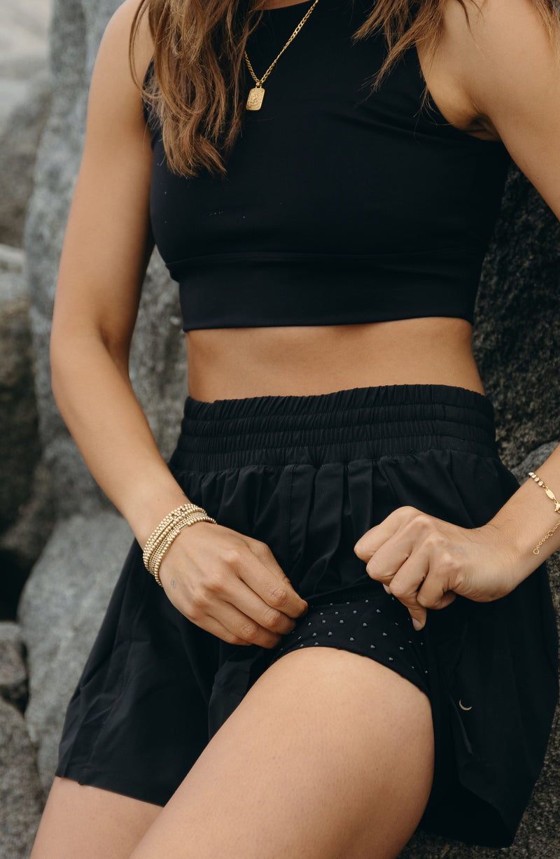Model wearing the L’Original Flowy Short in black with a featherlight pleated outer layer and sculpting compressive inner short. This 2-in-1 design combines feminine movement with performance support, featuring Elastique’s patented MicroPerle® bead infusion and a hidden inner pocket for essentials like a phone or tennis ball.