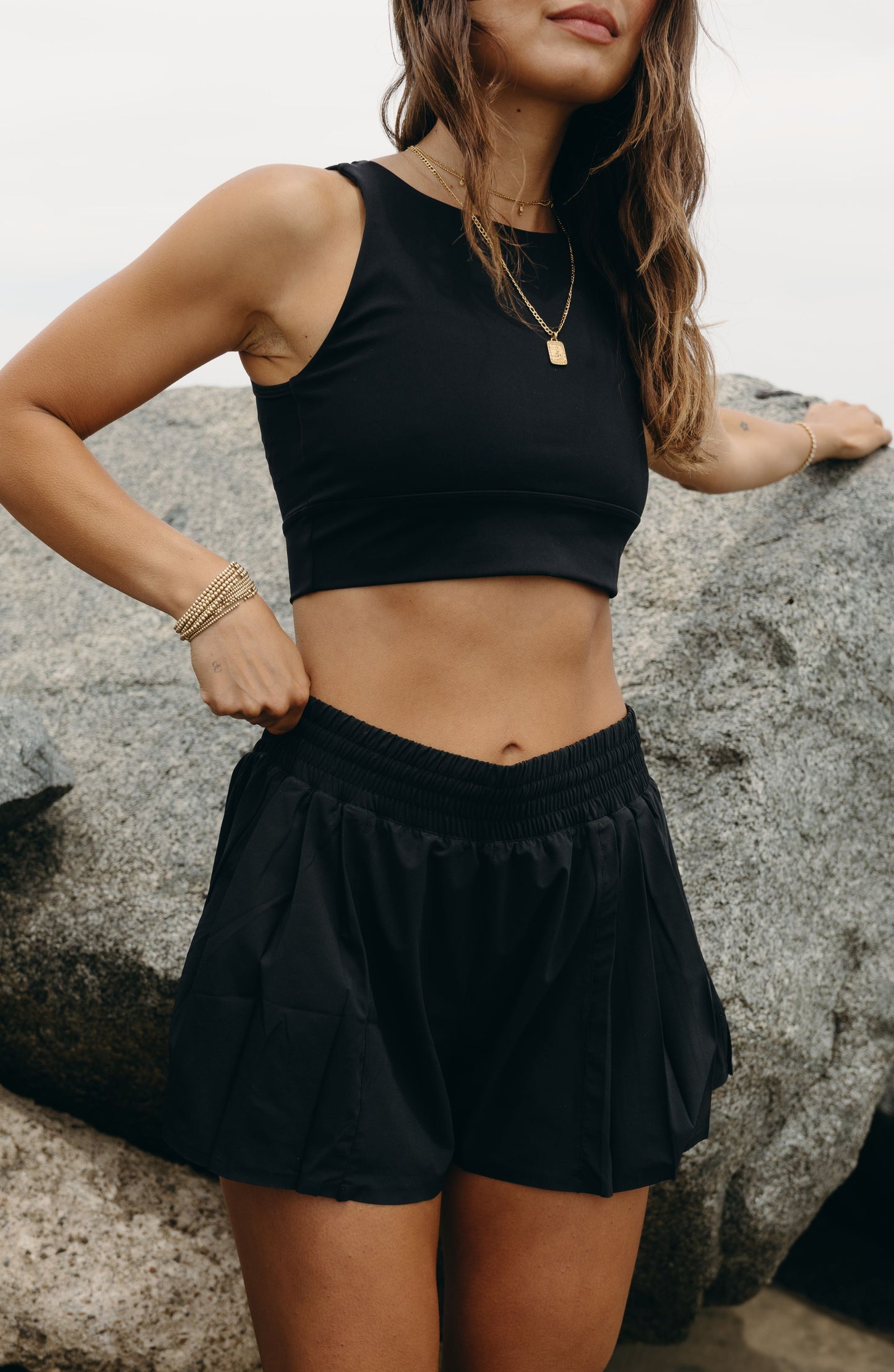 Model wearing the L’Original Flowy Short in black with a featherlight pleated outer layer and sculpting compressive inner short. This 2-in-1 design combines feminine movement with performance support, featuring Elastique’s patented MicroPerle® bead infusion and a hidden inner pocket for essentials like a phone or tennis ball.