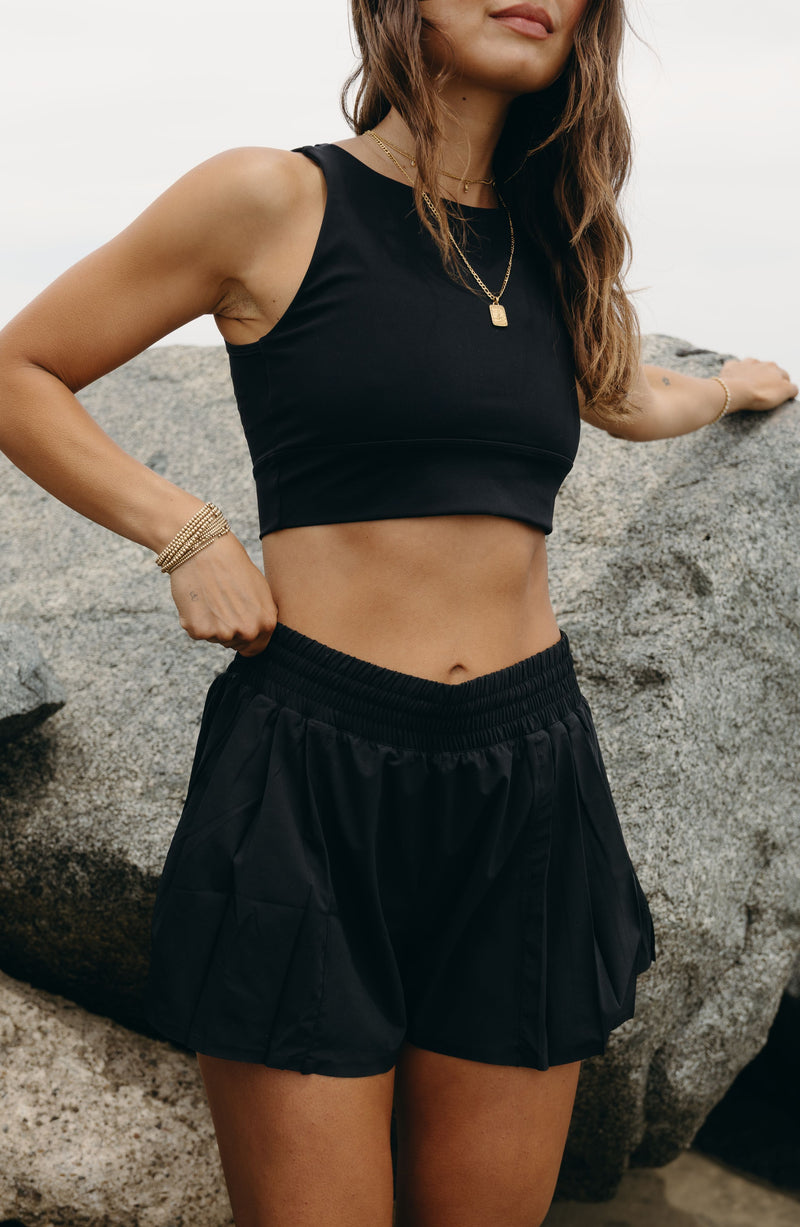 Model wearing the L’Original Flowy Short in black with a featherlight pleated outer layer and sculpting compressive inner short. This 2-in-1 design combines feminine movement with performance support, featuring Elastique’s patented MicroPerle® bead infusion and a hidden inner pocket for essentials like a phone or tennis ball.
