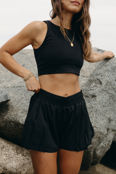 Model wearing L’Original Flowy Short featuring a featherlight pleated outer layer and a sculpting compressive inner short. The 2-in-1 design blends feminine movement with performance support, including patented MicroPerle® bead infusion and a hidden inner pocket for essentials like a phone or tennis ball.