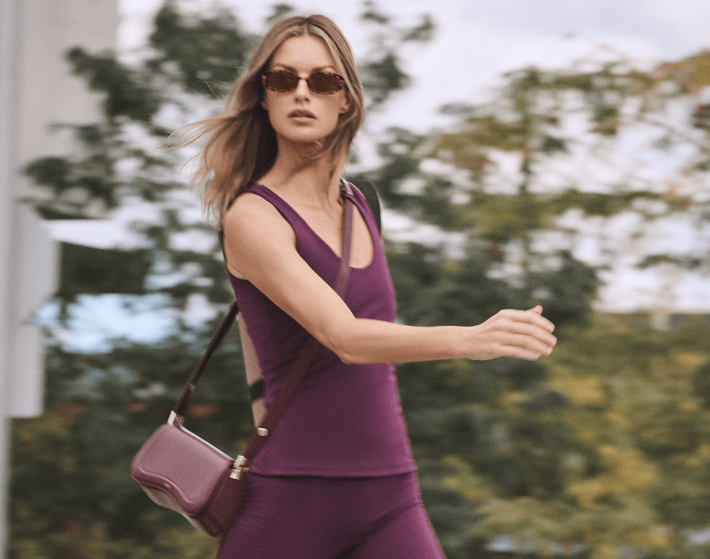 A woman wearing a matching purple activewear set and sunglasses walks outdoors while carrying a purple crossbody bag.