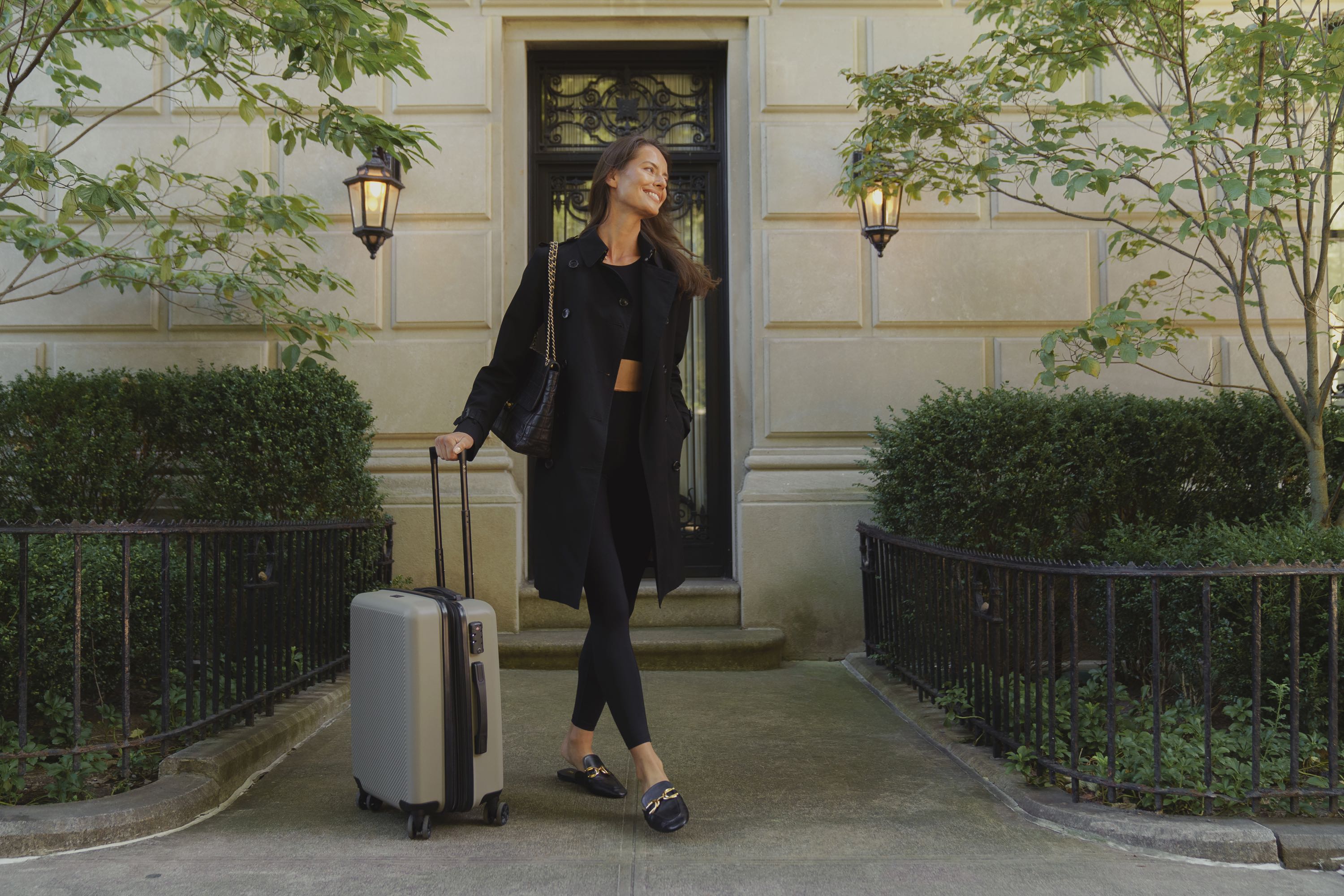 Women leaving town house with suite case for travel wearing black Elastique top and bottom along with trench coat, purse and slides.