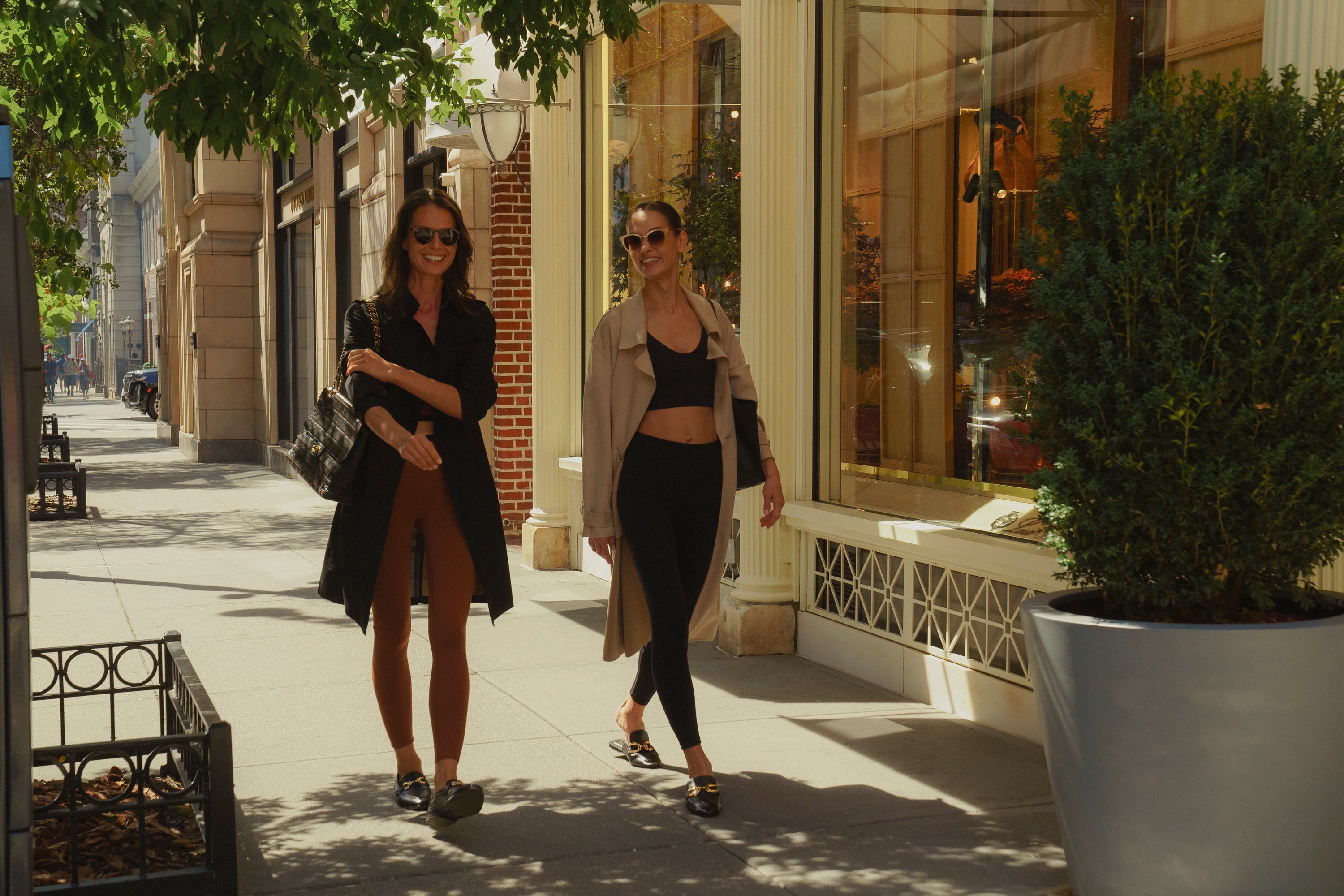 Two woemn walking down city street wearing Elastique tops and bottoms with trench coats sunglasses and purses. 