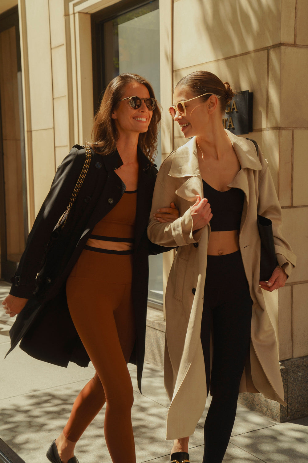 Two woemn walking down city street wearing Elastique tops and bottoms with trench coats sunglasses and purses. 