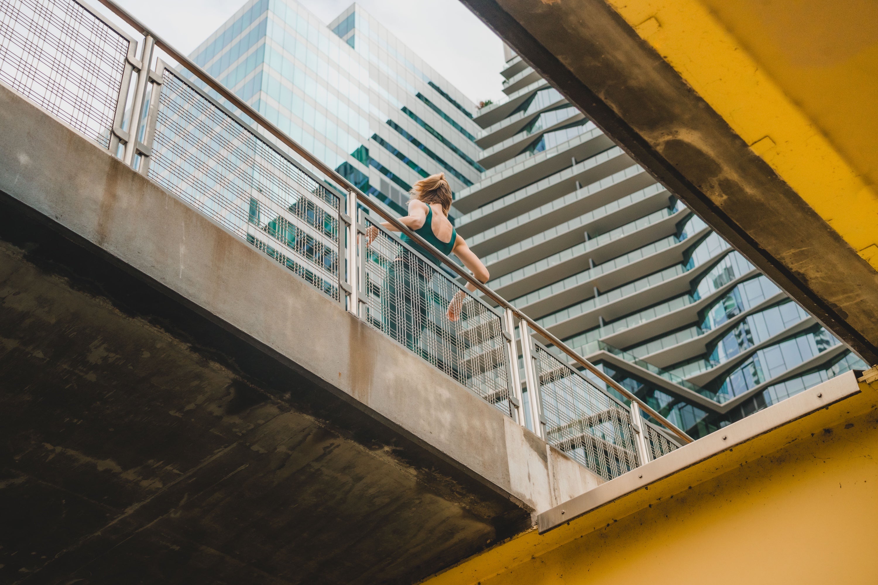 Image from a level down looking up to model leaning on railing in green leggings and bra top. City sky scape behind.