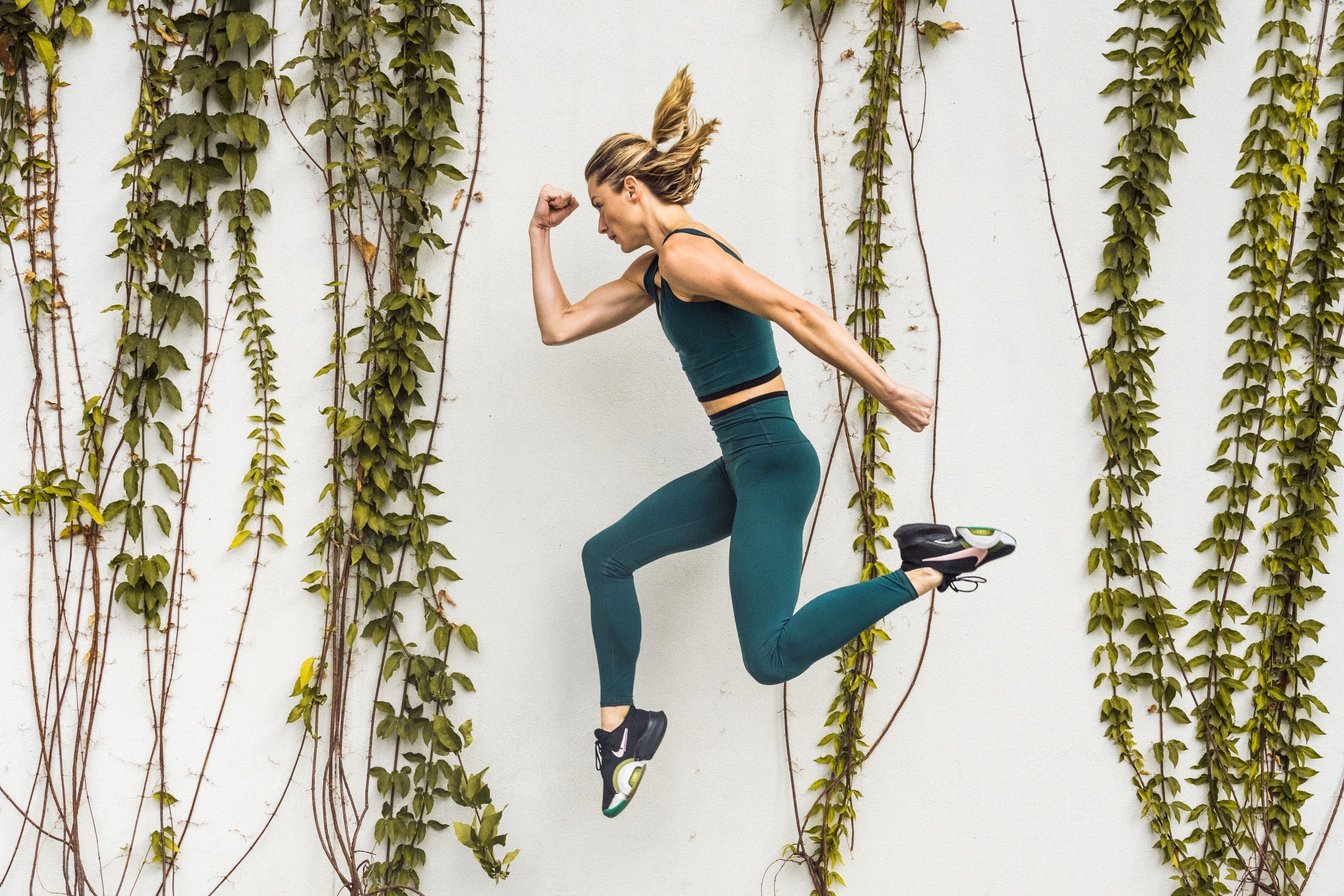 Women leaping through air in strong pose wearing green leggings and bra top and sneakers. Background is white wall with green ivy.