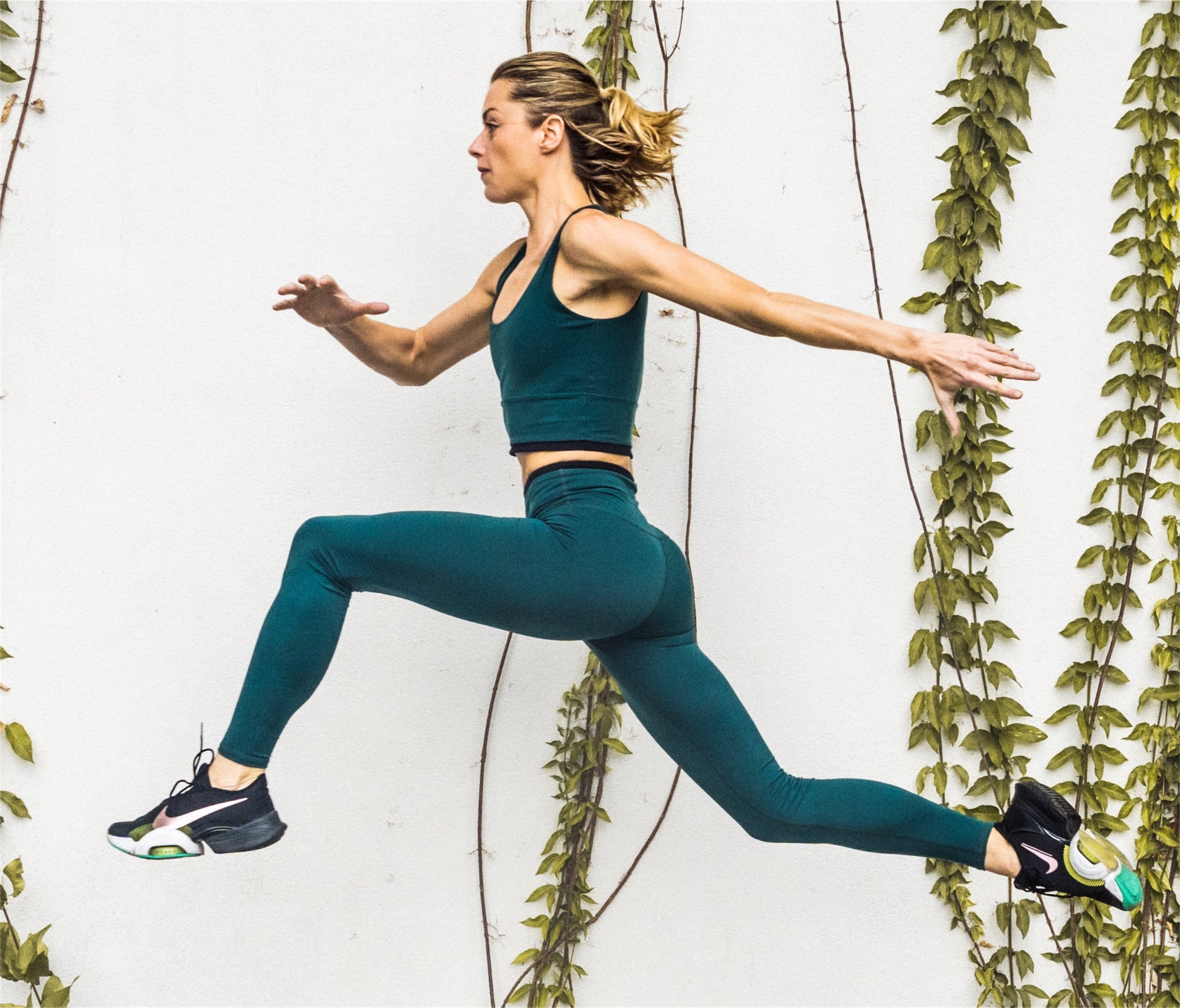 Women leaping through air in green leggings and bra top and sneakers. Background is white wall with green ivy.