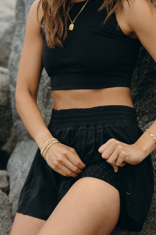 Load image 1 in gallery view. Model wearing the L’Original Flowy Short in black with a featherlight pleated outer layer and sculpting compressive inner short. This 2-in-1 design combines feminine movement with performance support, featuring Elastique’s patented MicroPerle® bead infusion and a hidden inner pocket for essentials like a phone or tennis ball.