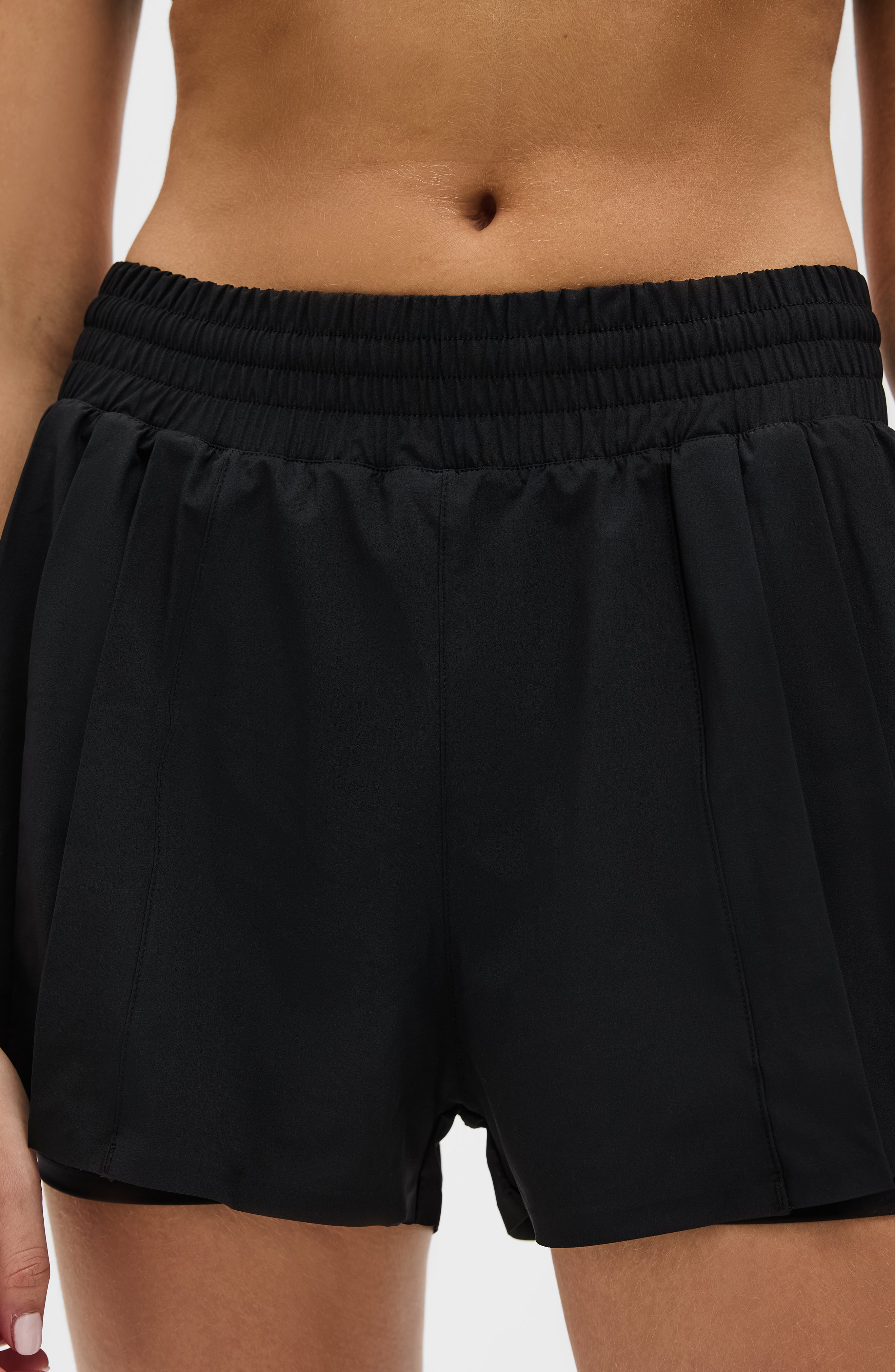 Model wearing the L’Original Flowy Short in black with a featherlight pleated outer layer and sculpting compressive inner short. This 2-in-1 design combines feminine movement with performance support, featuring Elastique’s patented MicroPerle® bead infusion and a hidden inner pocket for essentials like a phone or tennis ball.